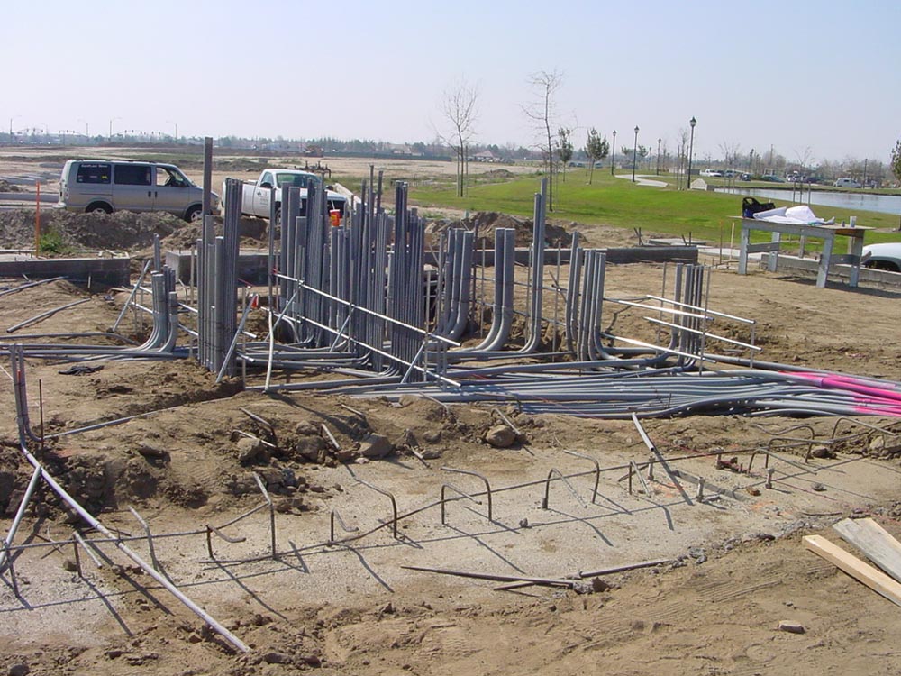 Construction site with clusters of gray electrical conduits emerging from dirt, parked vans and grassy park in background.