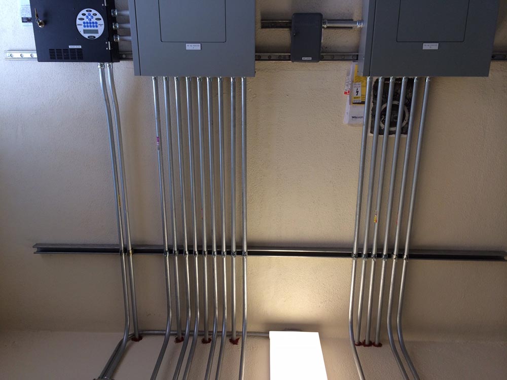 Ceiling view of metal electrical conduits neatly aligned feeding two gray electrical panels and a junction box