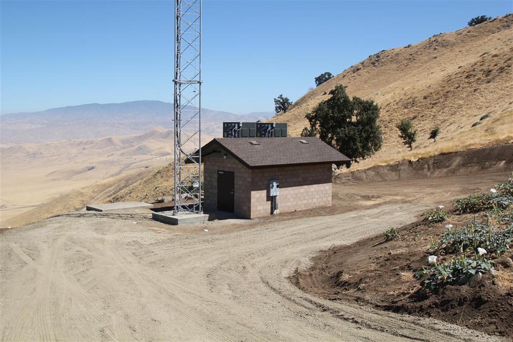 Telecom tower and small utility building beside a dirt road on an arid hillside with distant mountains