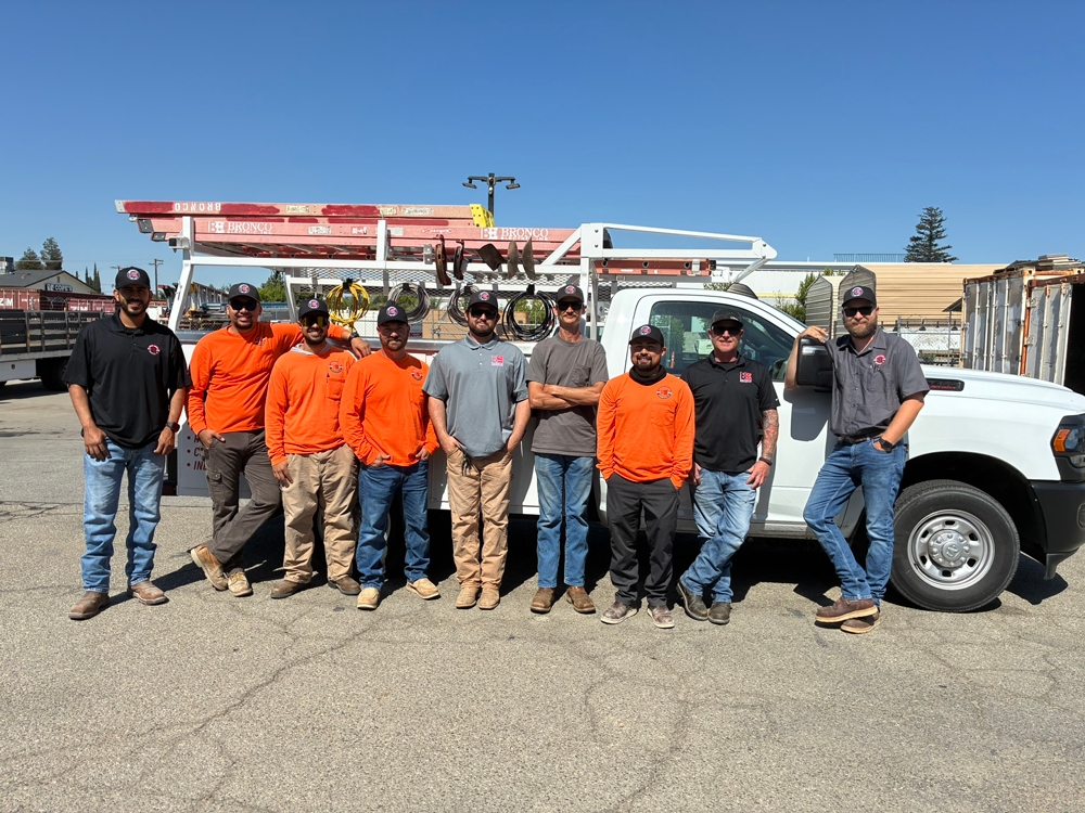 Construction crew of nine in orange work shirts posing by white utility truck with ladders under clear blue sky
