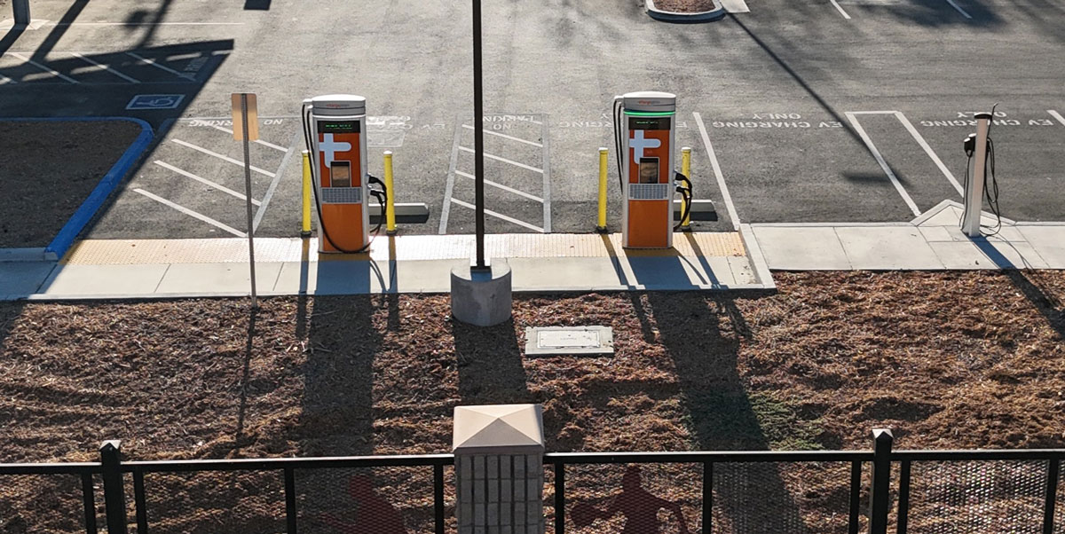 Two orange electric vehicle charging stations in an empty parking lot, parking spaces and long shadows visible.