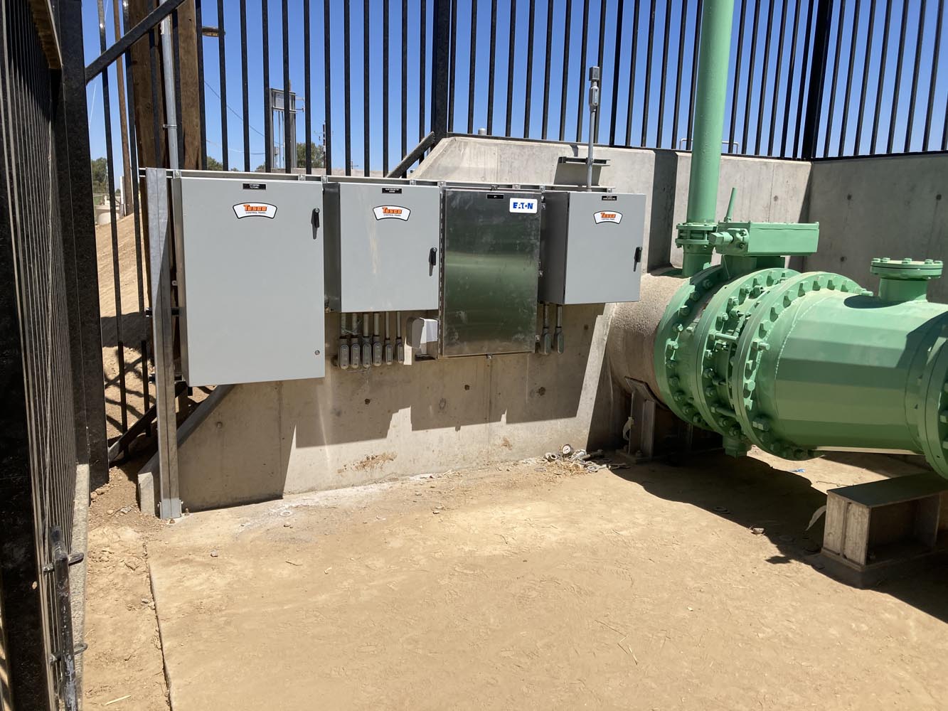 Outdoor fenced utility area with electrical control panels mounted on concrete beside large green pipeline valve