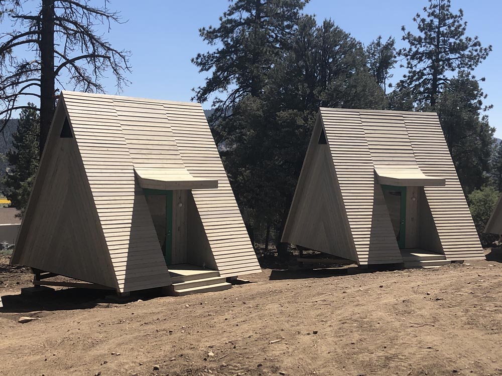 Two A-frame wooden cabins with green doors on a dirt clearing by pine trees under a clear blue sky