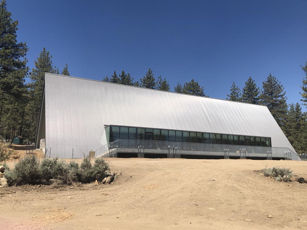 Large modern silver A-frame building with floor-to-ceiling windows in pine forest clearing, dirt foreground
