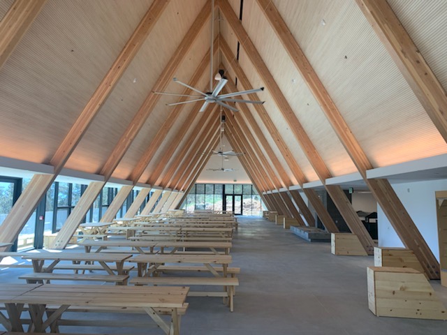 Spacious A-frame dining hall with exposed wooden beams, long picnic tables, ceiling fans, and large windows