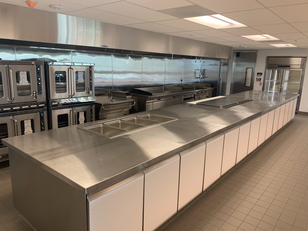 Stainless-steel commercial kitchen with long prep island, food wells, multiple ovens, range and undercounter cabinets