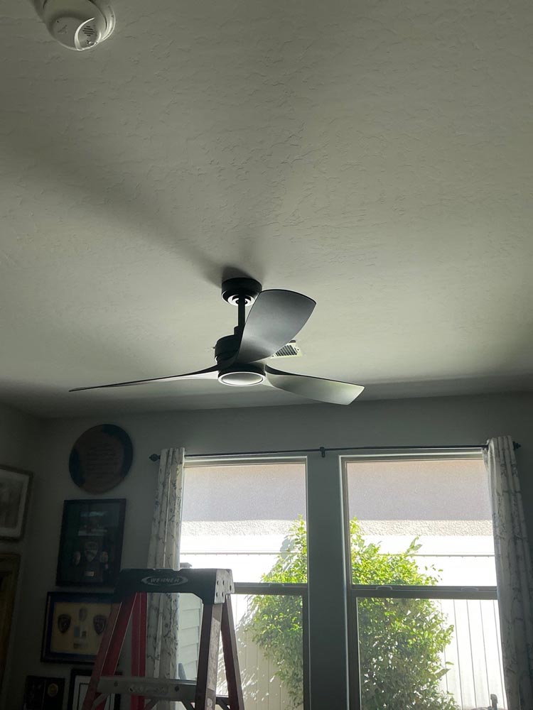 Modern black three-blade ceiling fan mounted on living room ceiling above ladder near windows and framed wall decor